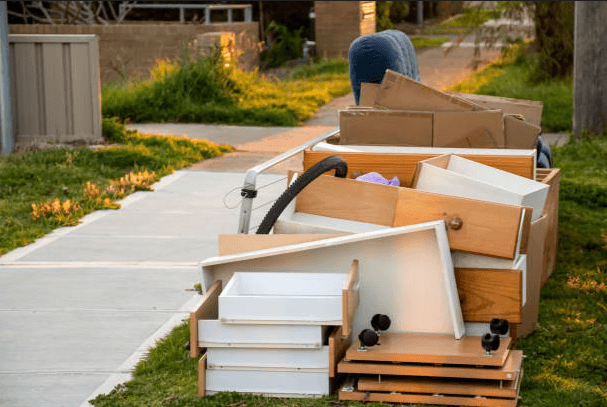 Discarded wooden furniture, white drawers, and cardboard boxes piled on a grassy suburban curb.